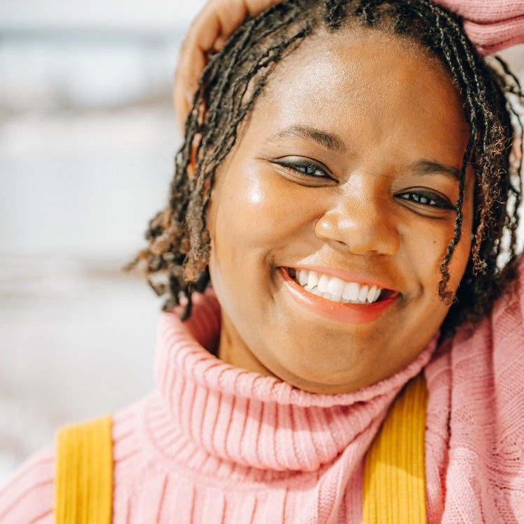 A Black person with braids, wearing a pale pink sweater and mustard overalls.
