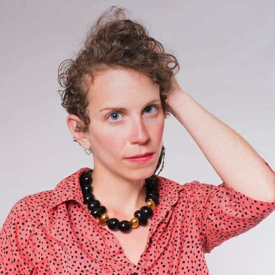 A white person with short, curly, dark blonde hair and a serious expression, wearing a black beaded necklace and black polka-dotted coral button down shirt.
