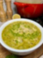 Bowl of soup with pesto and parmesan, set on a wooden board. Red pot, wooden shakers, lemon, and stove in the background. Cozy setting.