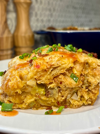 Close-up of breakfast casserole topped with green onions and hot sauce on a white plate. Background shows a cutting board and spice jars.