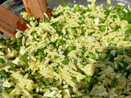 A bowl of pasta salad outside with orzo, spinach, peas and feta. Two salad tossers are in the bowl. and you can see the pool in the background. Bright, spring and summer vibes.