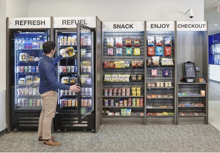 A person choosing a drink from an open-shelf micro market with cooler doors, snack shelves and a checkout kiosk