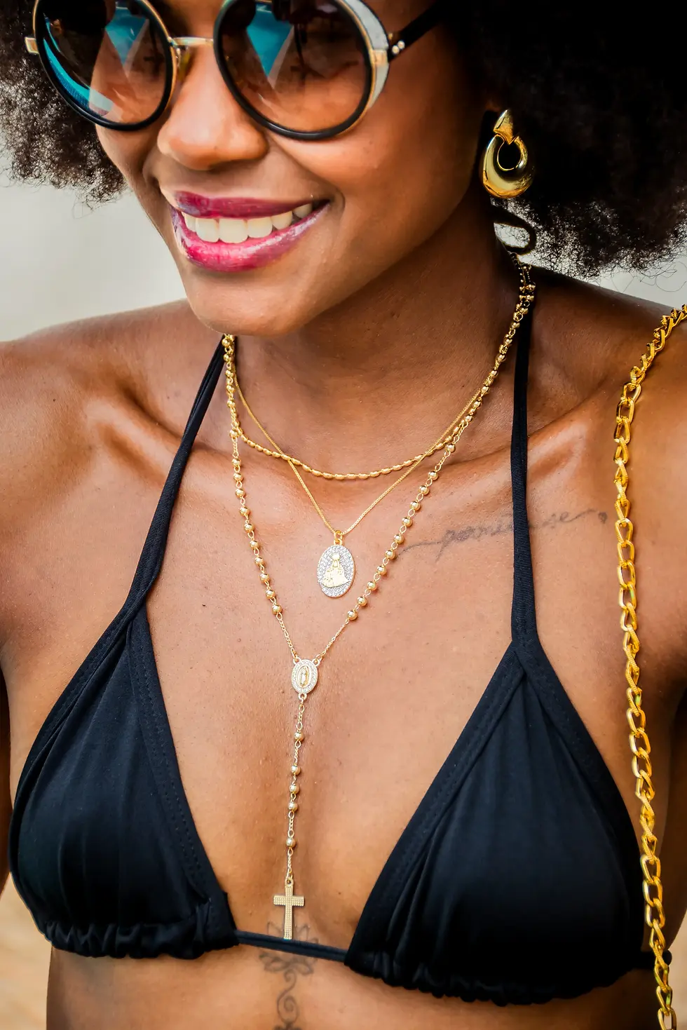 Ela veste um mix de três colares dourados Lèontinte sobre a pele iluminada, com destaque para o Colar Terço Longo de Nossa Se