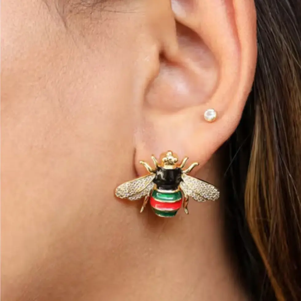 Na orelha, o Brinco Abelha Tricolor Lèontinte ganha destaque com suas listras em verde e vermelho e asas brilhantes.