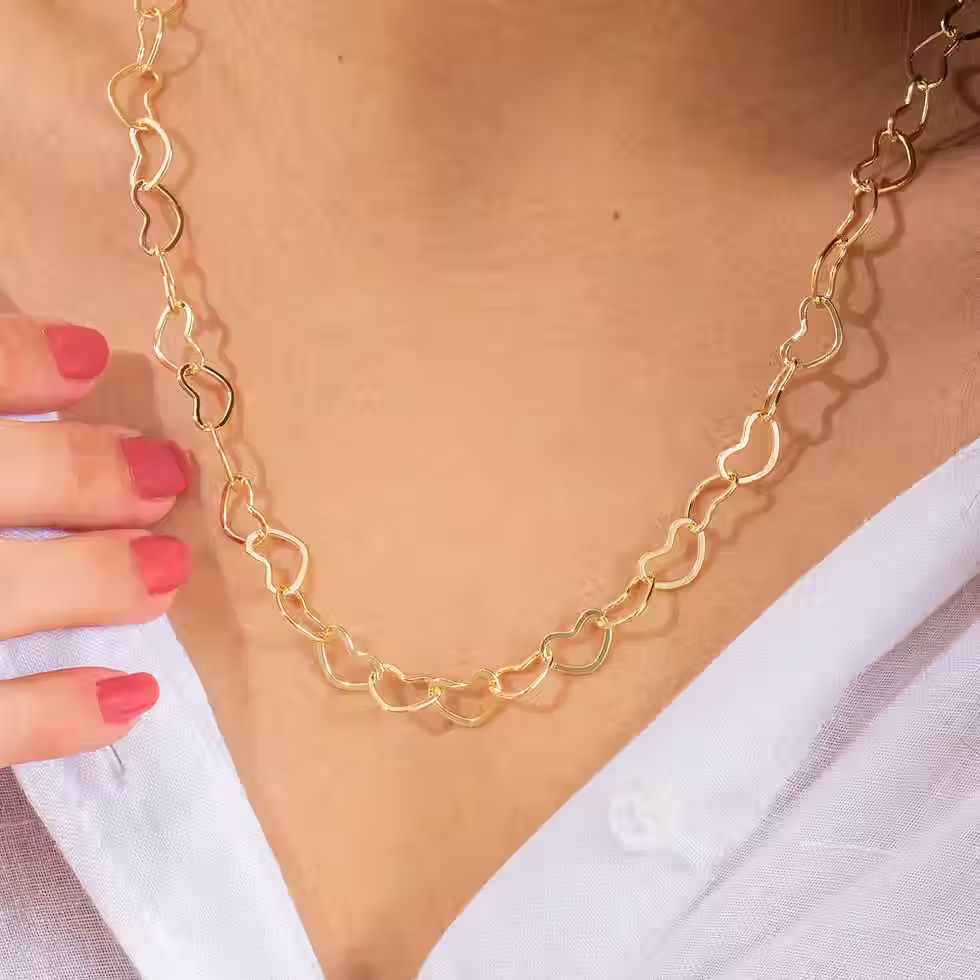 Detalhe do colo de uma modelo usando o Colar de Elos em Coração dourado com camisa branca, evidenciando o design delicado e r