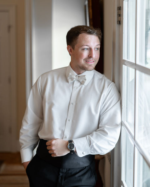 Groom adjusts his bow tie and cufflinks by a window while getting ready at Oak Island Mansion