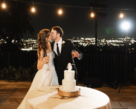 Bride and groom sharing a kiss while holding champagne glasses beside their wedding cake, with a beautiful evening city view at Vulcan Park and Museum in Birmingham, Alabama