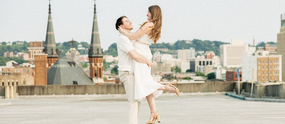 Newly engaged couple twirling on a Birmingham rooftop, he joyfully holds her in his arms as they smile and look at each other with the city skyline beautifully visible in the background.