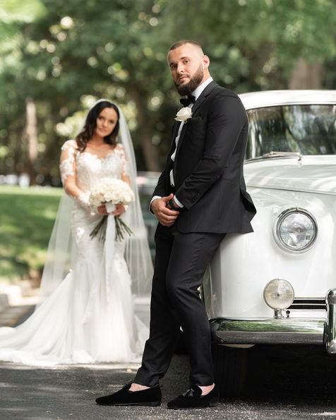 Editorial black-tie groom portrait leaning on a vintage white luxury wedding car with the bride framing the background, photographed by Kubay Studio.