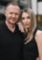 Outdoor portrait of Walter from Kubay Studio standing beside his wife, both wearing black outfits and smiling with modern city buildings softly blurred in the background.