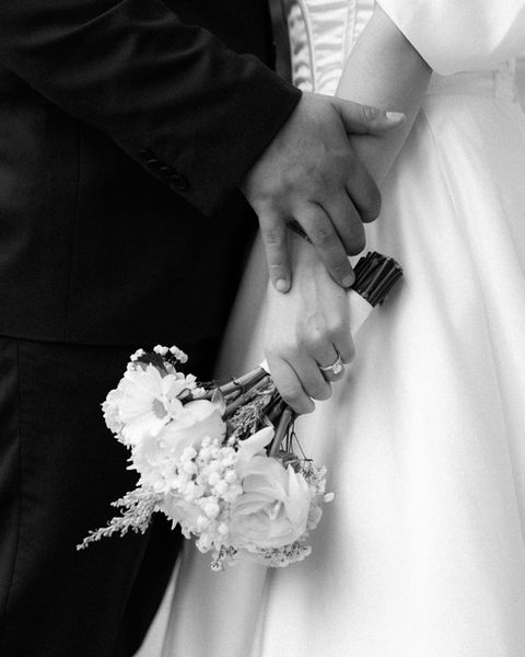 Close-up of bride and groom's hands holding bouquet with white flowers and rings visible.