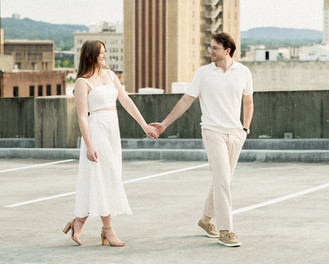 Newly engaged couple walking hand in hand across a rooftop in downtown Birmingham, Alabama, gazing at each other with the city skyline beautifully spread out behind them.