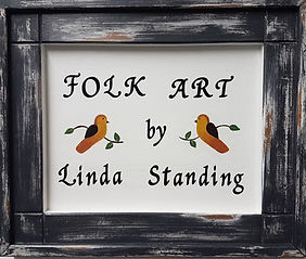 Black picture frame with title inside saying folk art by Linda Standing, decorated with two orange and black birds sitting on tiny twigs.