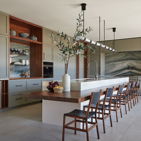 Modern kitchen with gray cabinets, marble backsplash, and long island. Wooden table, chairs, and fruit basket. Vase with branches. Minimalist decor. GRAY Awards. GRAY magazine.