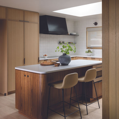 Modern kitchen with wood cabinets, an island with stools, and a skylight. A green plant in a vase and bread on the counter add warmth. GRAY Awards. GRAY magazine.