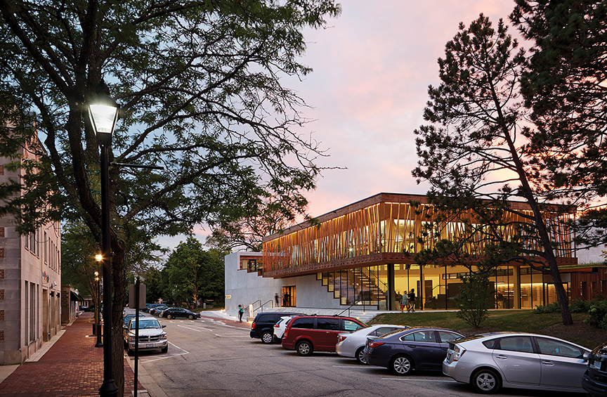 Writers Theatre building in Glencoe, Illinois, at dusk with pink clouds, tree-lined street with parked cars, wood slat architecture, floor to ceiling glass windows, large staircase, concrete and wood