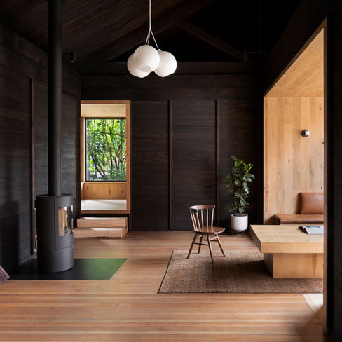 Cozy wood-paneled room with vintage chair, rectangular table, and a potted plant. Black stove, bright window, and modern light fixture. 