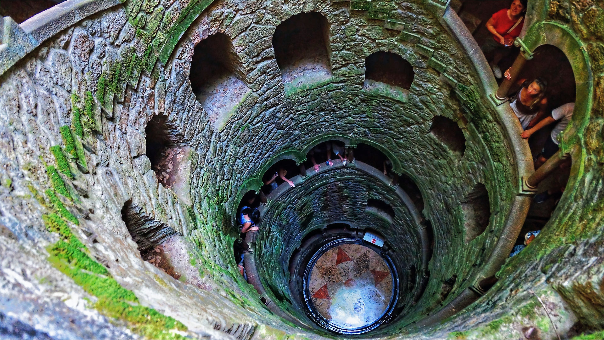 Wendeltreppe von oben fotografiert, sodass der Boden zu sehen ist. DIe Treppe ist durch eine Wand als Geländer getrennt, die voller Fenster ist.