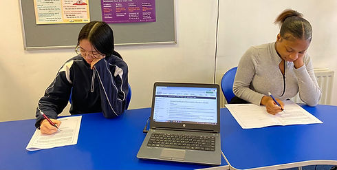 Two female students sitting at a blue desk taking notes beside an open laptop computer