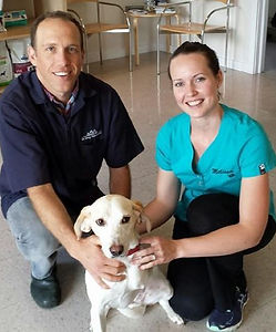 Two veterinary staff smile with a dog in a vet clinic setting.