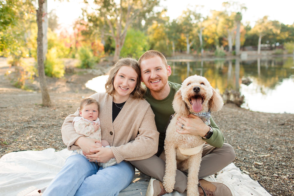 A couple smiles at the camera, holding a baby and a dog by a lake. Autumn trees and bright colors create a warm, happy scene. Late summer outdoor newborn session at Vasona Lake Park in Los Gatos by San Jose Bay Area Photographers HKCreate