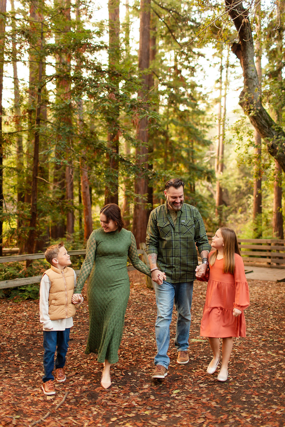 Family of four walking hand in hand in a forest. Parents and two children smiling. Green, brown, and orange clothing complement the autumn setting.
Fall family photoshoot at the Redwood Grove Nature Preserve in Los Altos by Bay Area Photographers HKCreate.