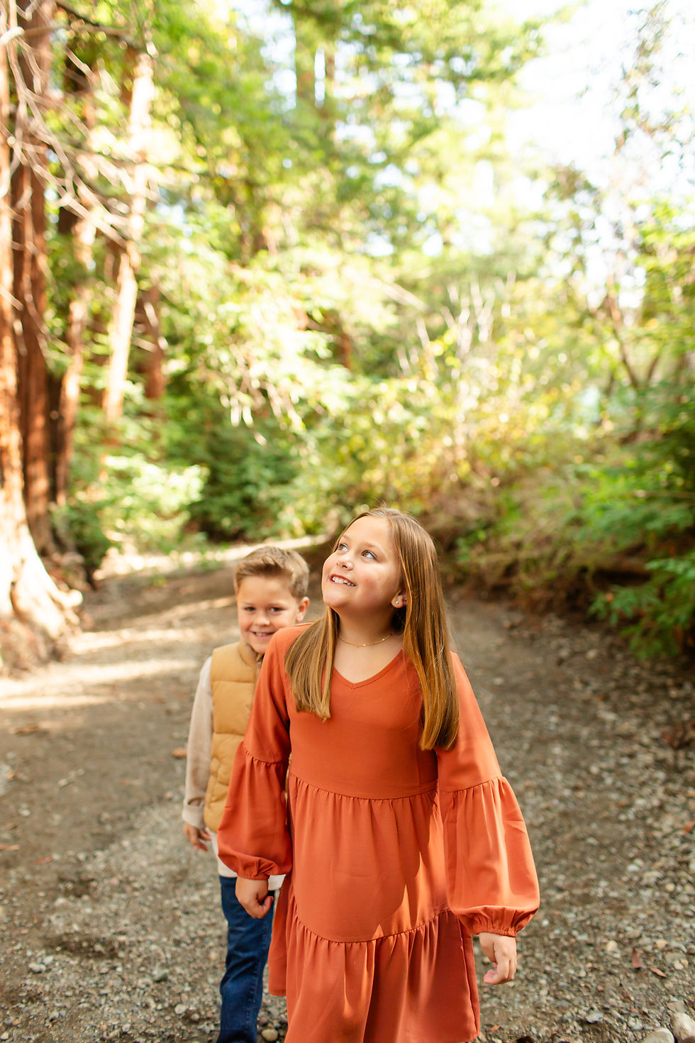 Two children walk in a forest, girl in orange dress smiling upwards, boy in beige vest. Sunlight filters through green trees, creating a cheerful mood.
Fall family photoshoot at the Redwood Grove Nature Preserve in Los Altos by Bay Area Photographers HKCreate.