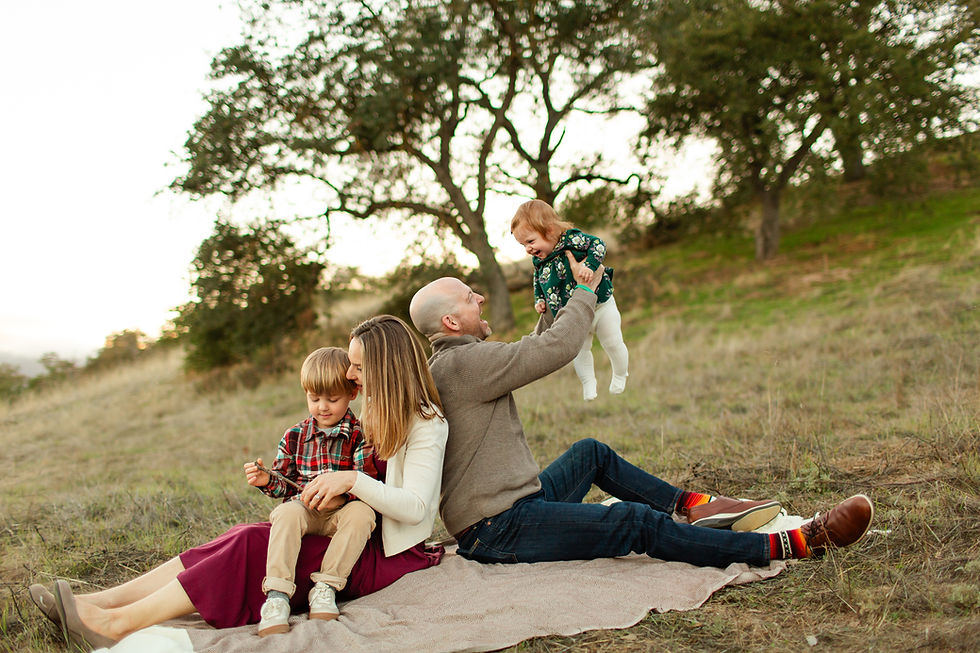 Family sitting on a blanket in a grassy field. A man lifts a child joyfully, while a woman cuddles another child. Trees in the background. Candid family moment interacting with each other.
Guadalupe Oak Grove Park in San Jose Winter Family of Four Photo Session during sunset by HKCreate Bay Area Photographers