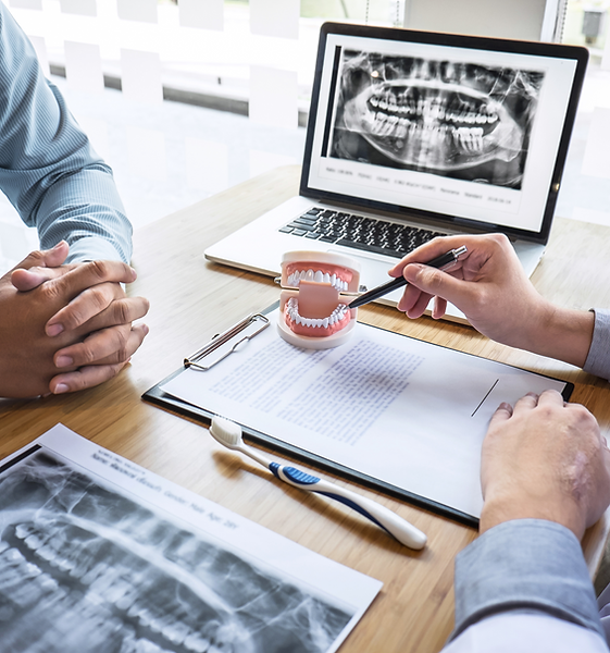 A dental consultation scene showing two people seated at a desk. On the desk are dental X-rays, a clipboard with documents, a toothbrush, a dental model of human teeth, and a pen. One person points to the model with the pen, while a laptop in the background displays a panoramic dental X-ray.