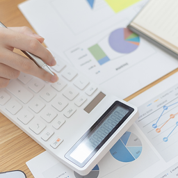 Close-up of a hand using a white calculator with a silver pen, surrounded by printed financial reports featuring pie charts, bar graphs, and a notebook. The image conveys data analysis, budgeting, or financial planning in a professional setting.