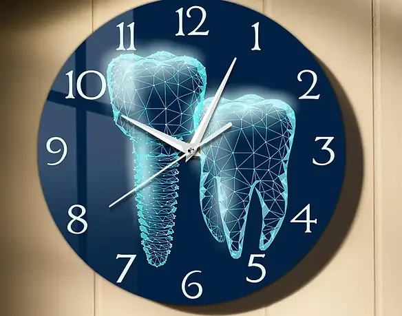 Wall clock with a dark blue face featuring white hour numbers and three clock hands. The center of the clock displays two glowing, wireframe-style 3D dental illustrations—one of a tooth and the other of a dental implant. The clock is tilted within a red border and mounted on a beige wall.