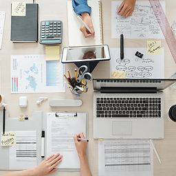 Top-down view of a busy workspace with multiple hands working on strategy and planning documents. The desk is filled with a laptop, tablet, calculator, charts, flowcharts, printed documents, sticky notes, pens, and office supplies. One note reads “deadline Monday.” The scene conveys active collaboration and project planning.
