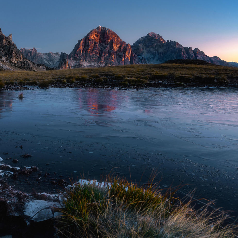 Sunlit mountain peaks отражаются in a frozen lake at dawn, with grassy foreground and a clear blue-pink sky.