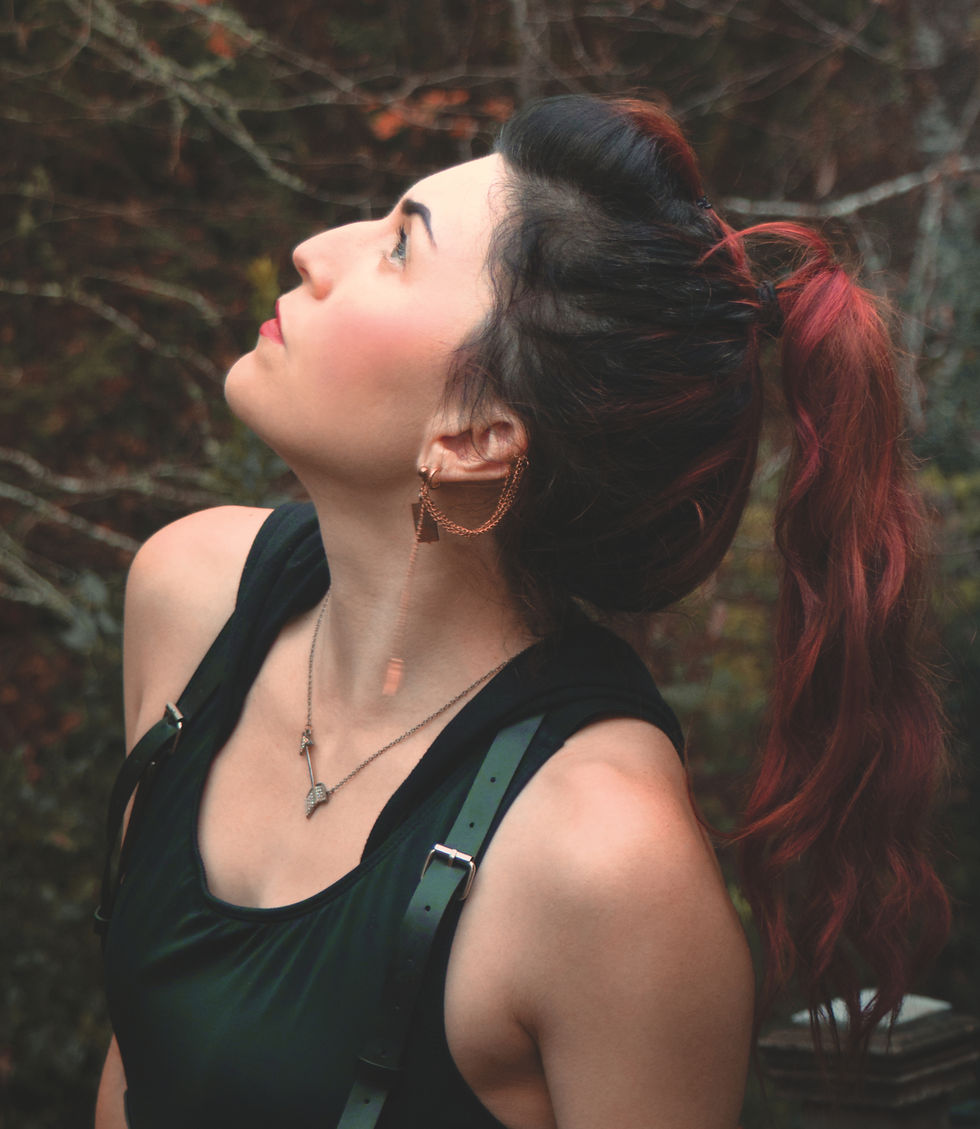 Woman with red hair in ponytail looks up, wearing black top and earrings, against a blurred forest background. Warm, contemplative mood.