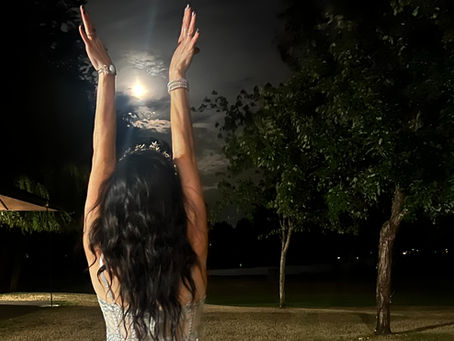 Woman in a silver gown stands on a grassy lawn at night, arms raised toward the moonlit sky, with trees in the background.