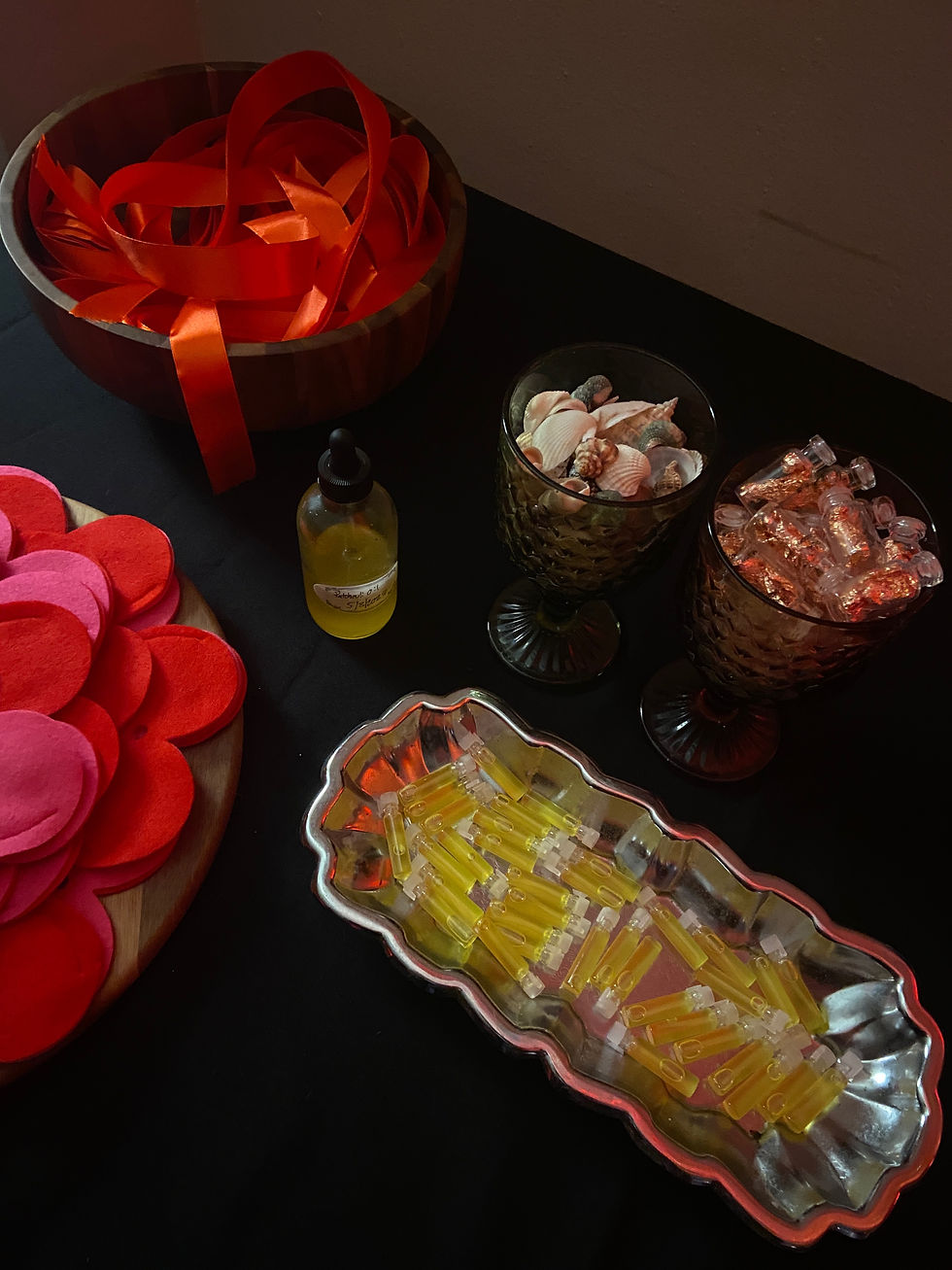 Wooden bowl with red ribbons, felt circles, oil bottle, shells, and tubes on a black table. Warm, crafty atmosphere.