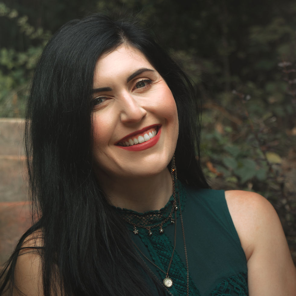 Smiling woman with long black hair in a dark green top and layered necklaces, posing outdoors against a blurred leafy background.