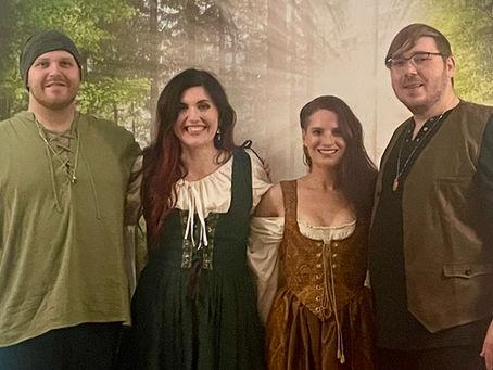 Four people in medieval costumes smile in front of a forest backdrop. Warm lighting and pink string lights create a festive atmosphere.