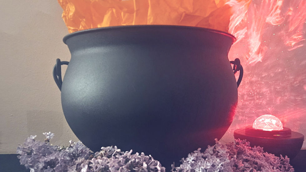 Black cauldron with orange paper flames, surrounded by lilac flowers on a black cloth. Red glow and light patterns in the background.