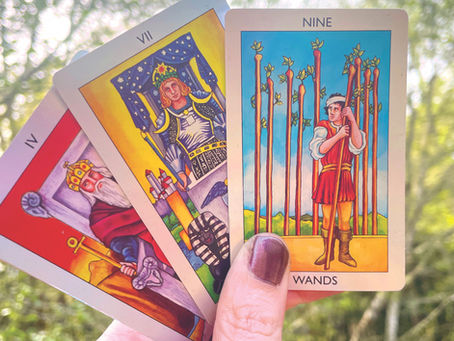 A hand holds three colorful tarot cards, featuring figures and symbols, against a blurry green forest background.