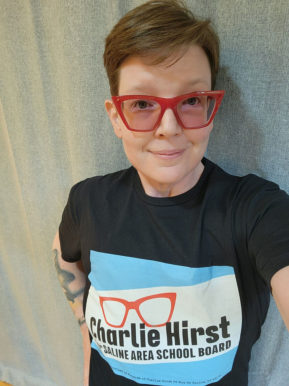 Image description: A white person with red glasses wearing a black short-sleeve t-shirt that has Charlie's campaign logo. The logo has red eyeglasses above Charlie Hirst in black font and Saline Area School Board in blue font.