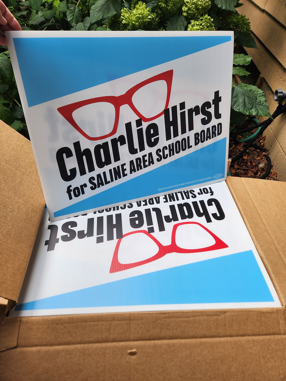 Image description: Campaign yard signs that read Charlie Hirst for Saline Area School Board " in black font with a pair of red eyeglasses and a blue and white background.