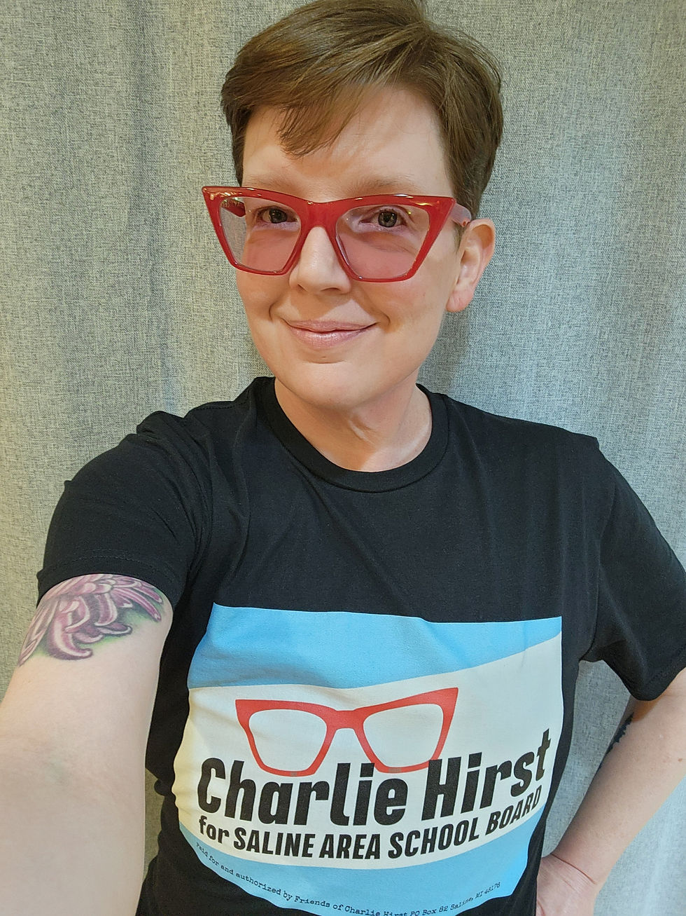 Image description: A white person with red glasses wearing a black short-sleeve t-shirt that has Charlie's campaign logo. The logo has red eyeglasses above Charlie Hirst in black font and Saline Area School Board in blue font.