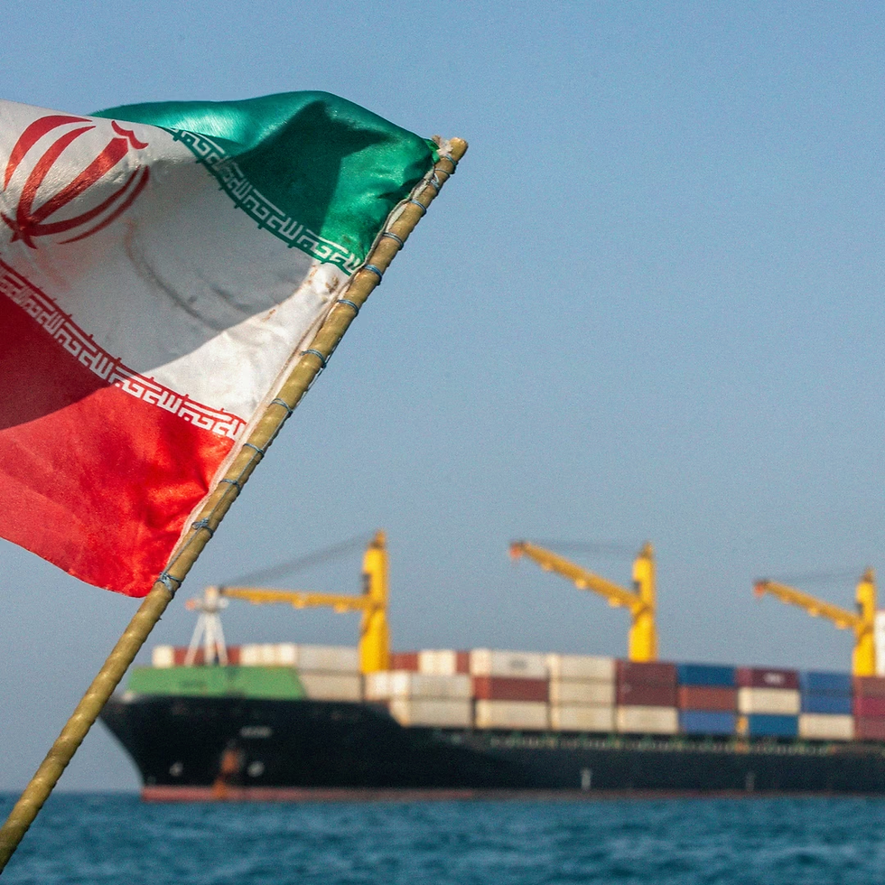Iranian flag in foreground with a container ship and yellow cranes on the sea under a hazy blue sky.