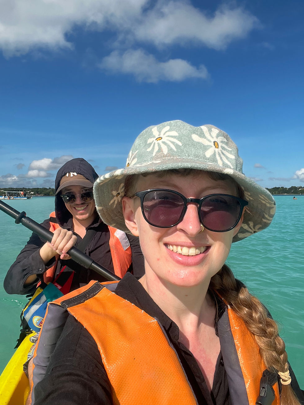 Two people kayaking on turquoise water under a blue sky. They wear orange life vests, sunglasses, and hats, smiling at the camera.