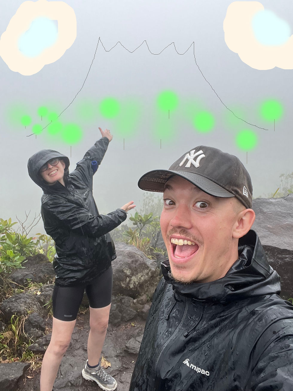 Two smiling hikers in rain gear on rocky trail, one pointing up at a fog-shrouded mountain outline in the misty background.