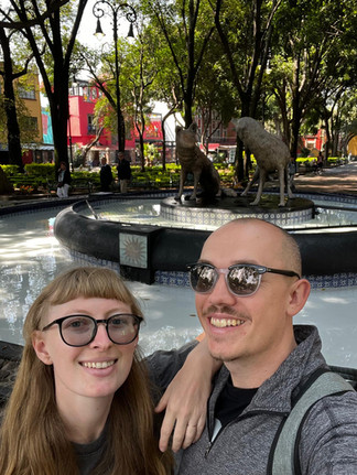a man and woman pose for a selfie in front of fountain showing statue of two coyotes.