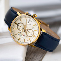 Gold-tone chronograph wristwatch with a cream dial and navy blue leather strap on a white fabric and wooden surface