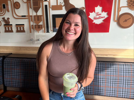 A woman sitting in a sunlit cafe booth, smiling, holding an iced matcha.