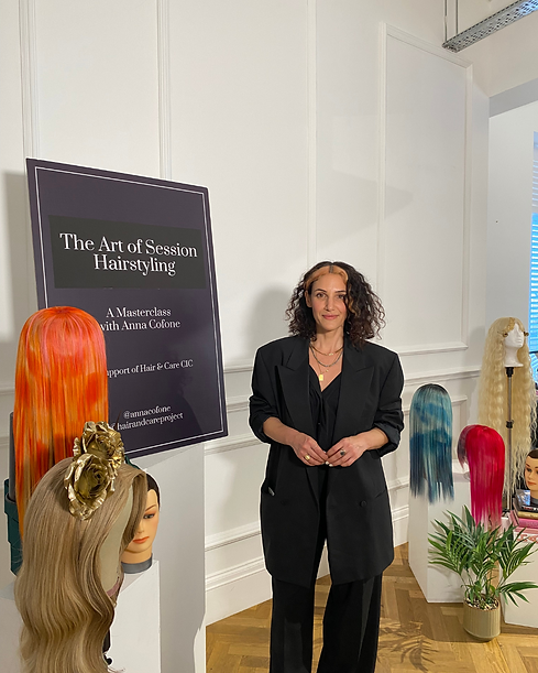 A photo of Anna, our founder, at 'The Art of Session Hairstyling' masterclass. She is stood in the middle with a selection of her wigs displayed around her. Behind her to the left is a dark banner with white writing, advertising 'The Art of Session Hairstyling'.